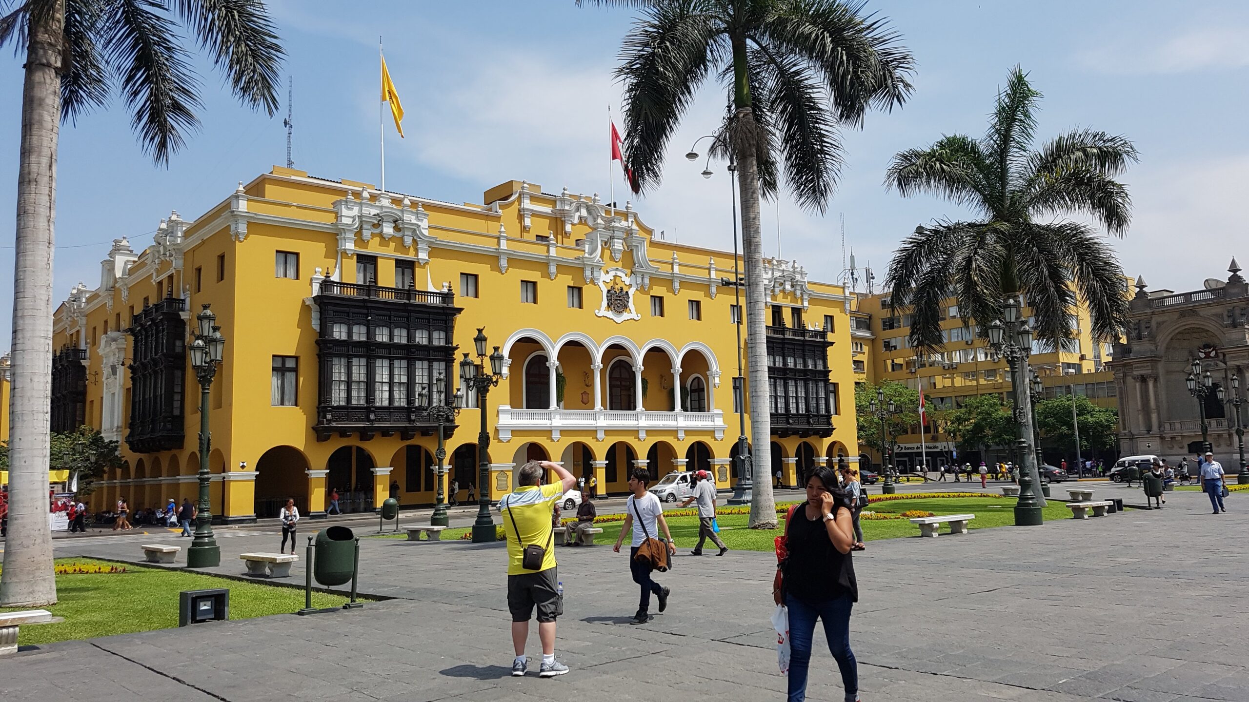 El Palacio Municipal de Lima, de fachada amarilla y balcones oscuros, se ve desde la plaza. Hay palmeras, banderas y gente paseando.