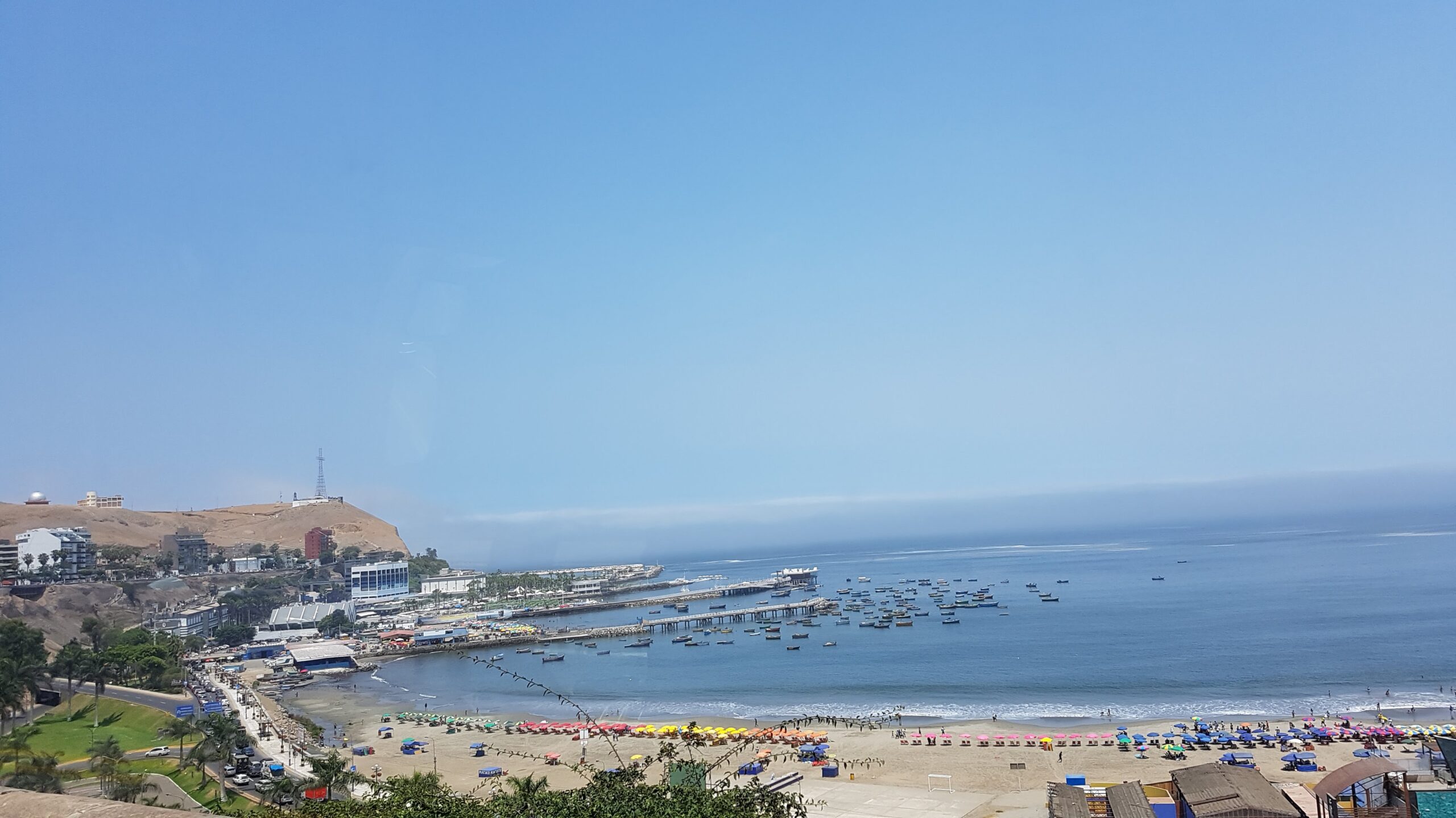 Una playa de Lima se ve desde lo alto, con sombrillas de colores y un muelle lleno de botes. El cerro baja hacia el mar bajo un cielo despejado.