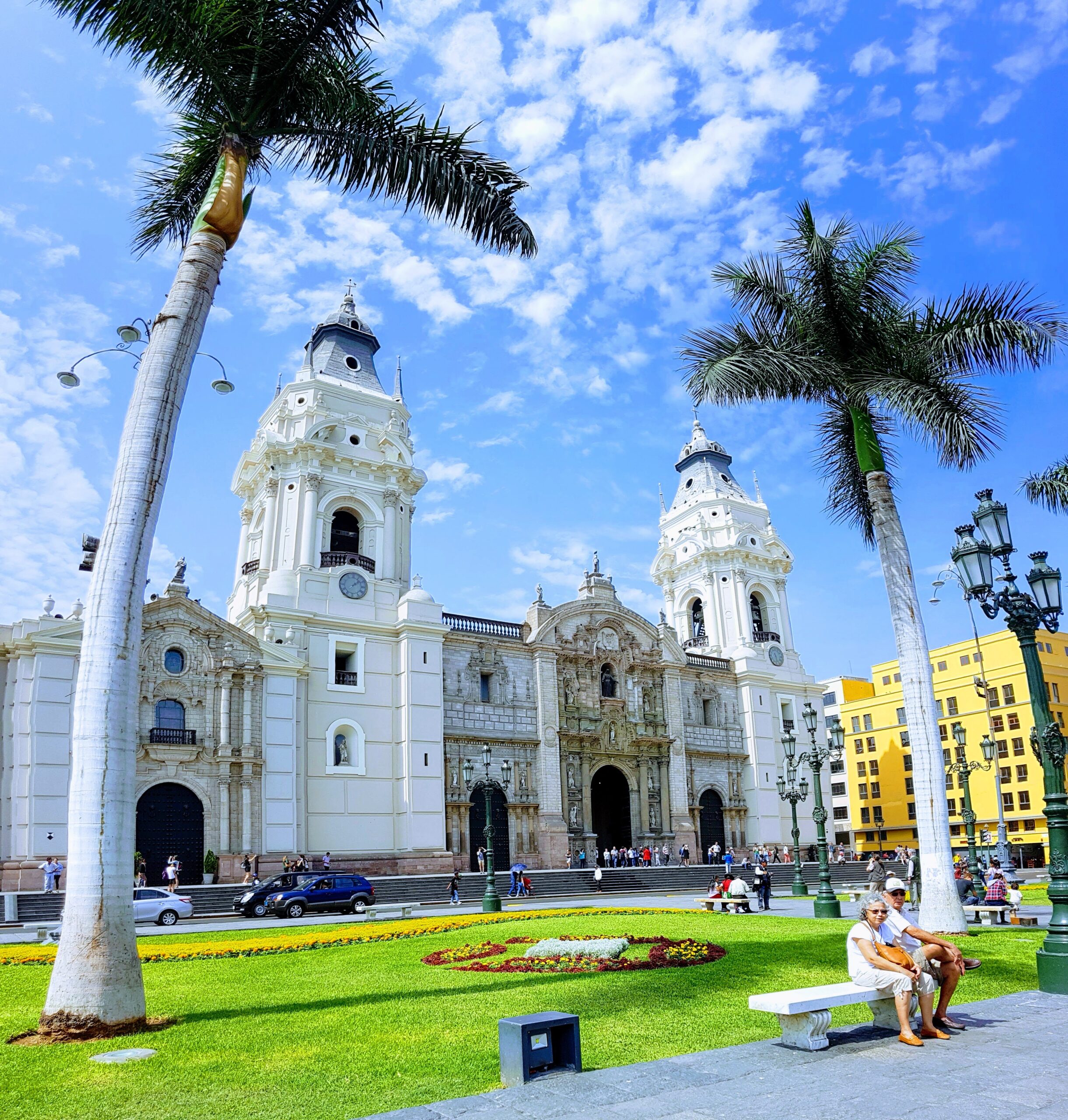 La Catedral de Lima se ve entre dos palmeras, con su fachada blanca y las dos torres. En el jardín hay flores y una pareja descansa en una banca.