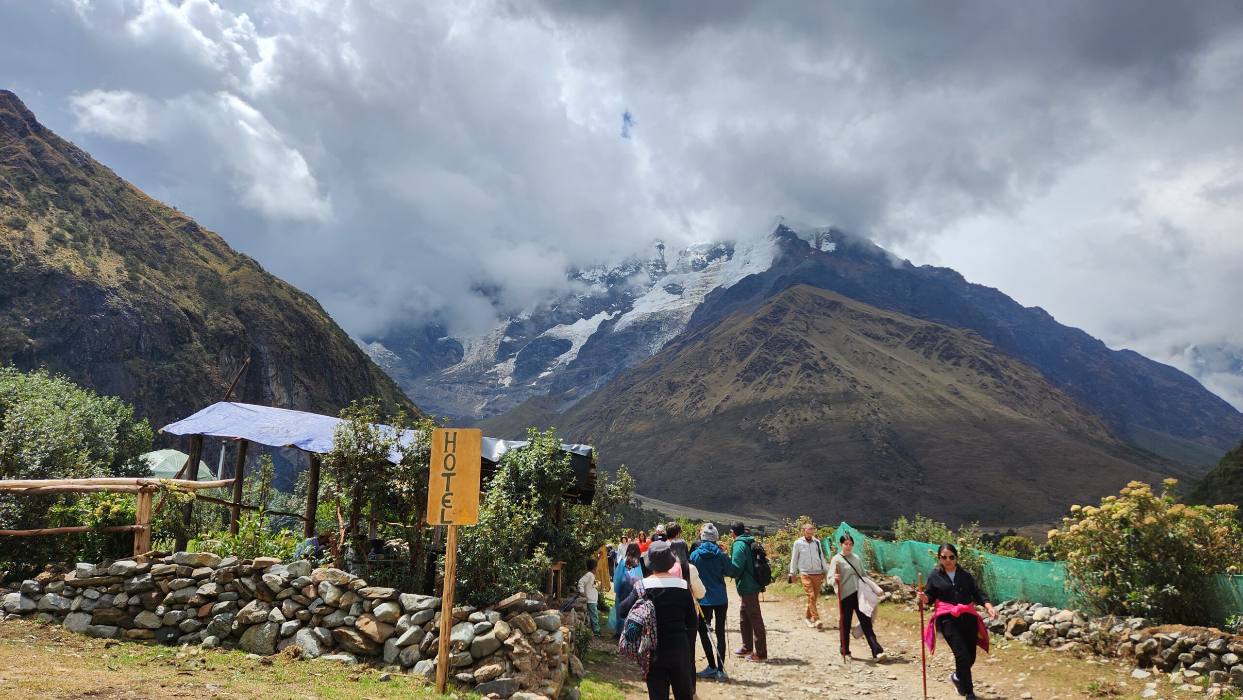 Un grupo camina por un sendero hacia la laguna Humantay, con el nevado entre las nubes. A un lado hay un muro de piedra y un letrero de hotel.