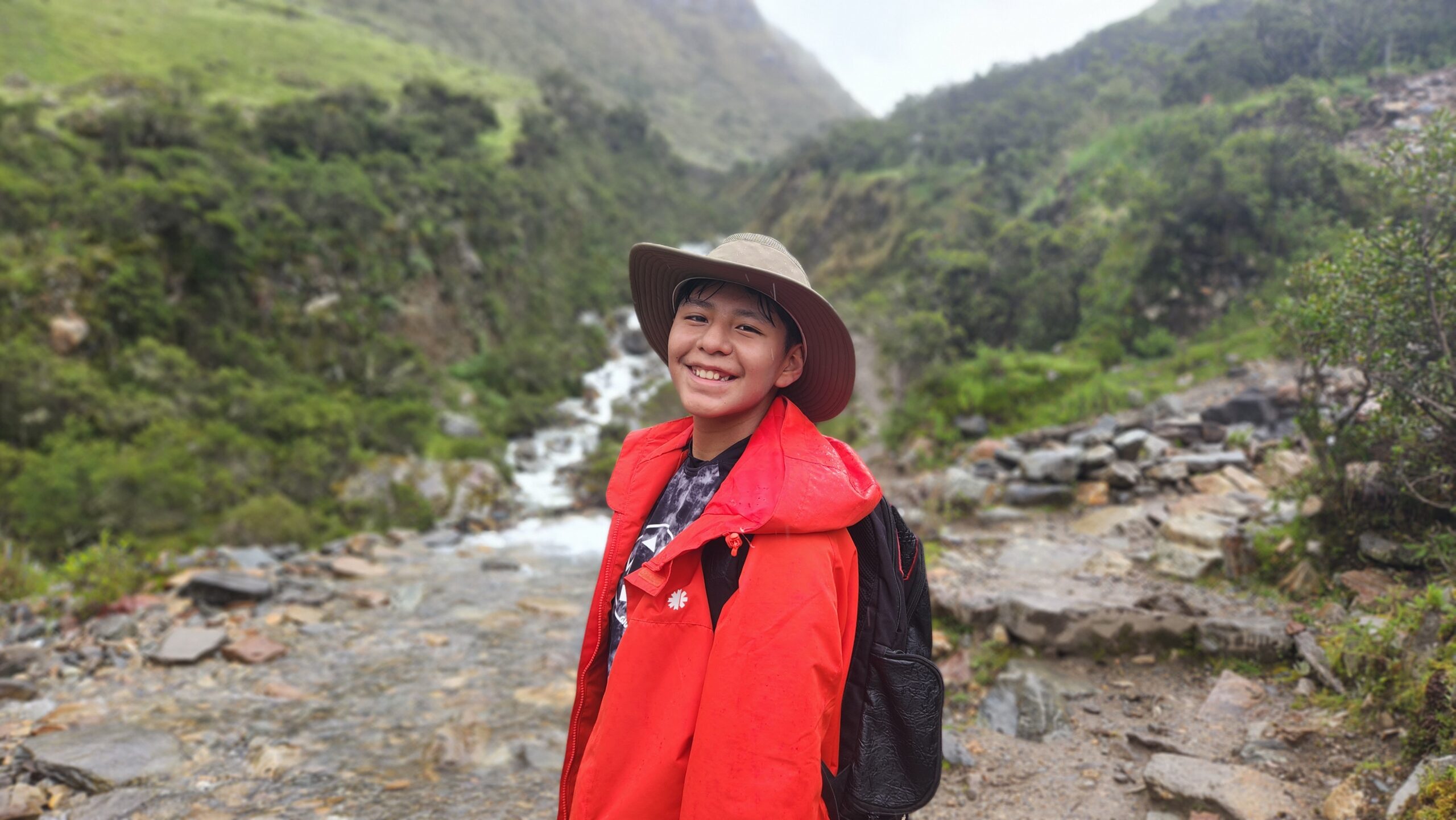 Un viajero con sombrero y chaqueta roja sonríe en un sendero de piedras. Detrás baja un riachuelo entre cerros verdes.
