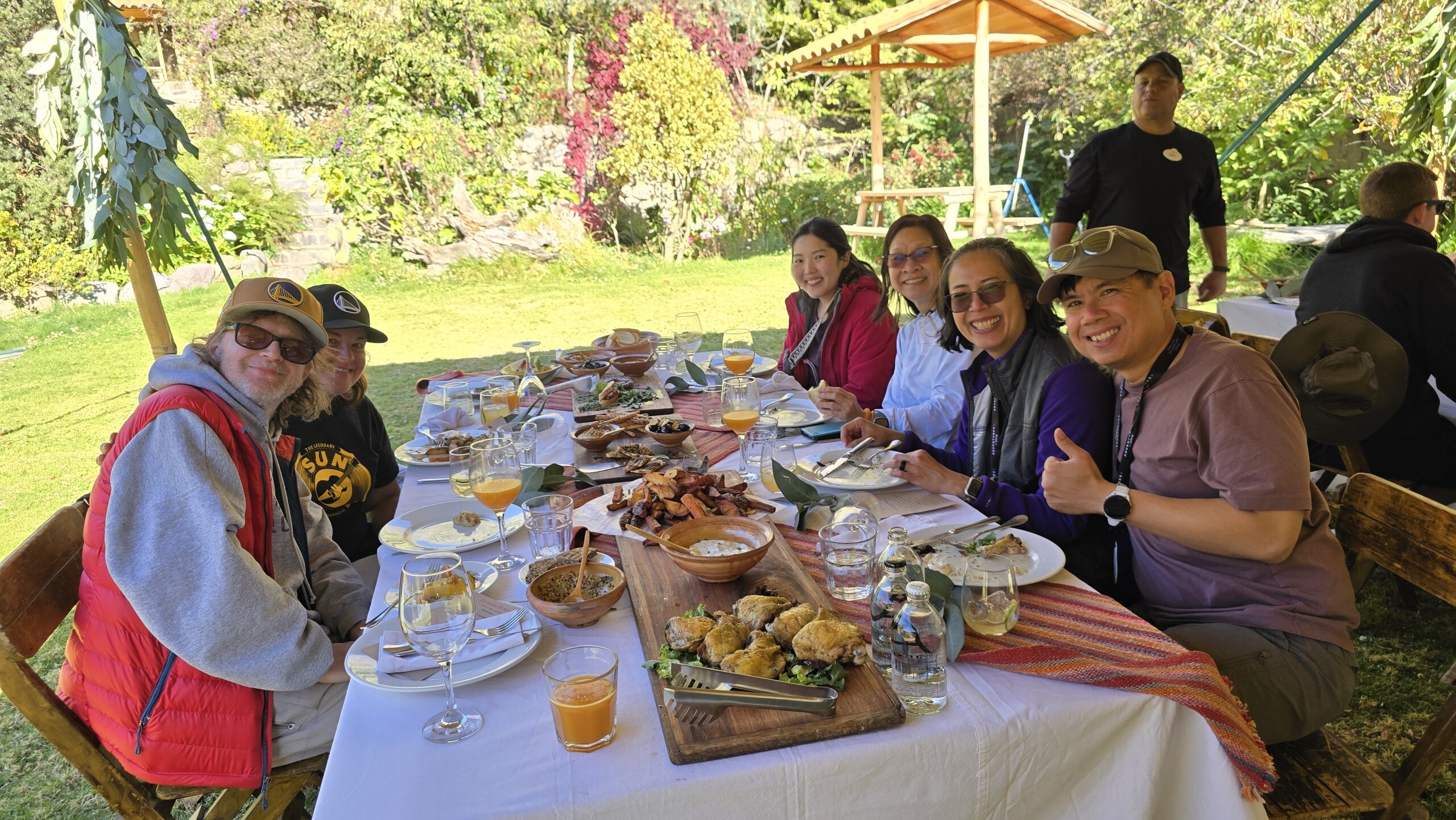 Un grupo almuerza en una mesa larga, en un jardín. Hay carne, jugos y platos de barro, con el cerro y las flores detrás.