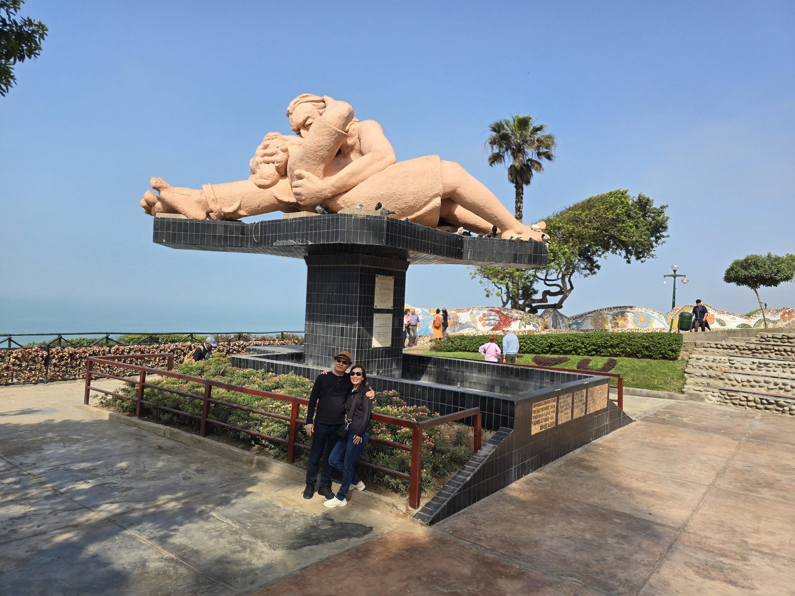 Una pareja sonríe frente a la estatua del Beso, en el Parque del Amor de Lima. Detrás se ve el mar, una palmera y el muro de mosaicos.