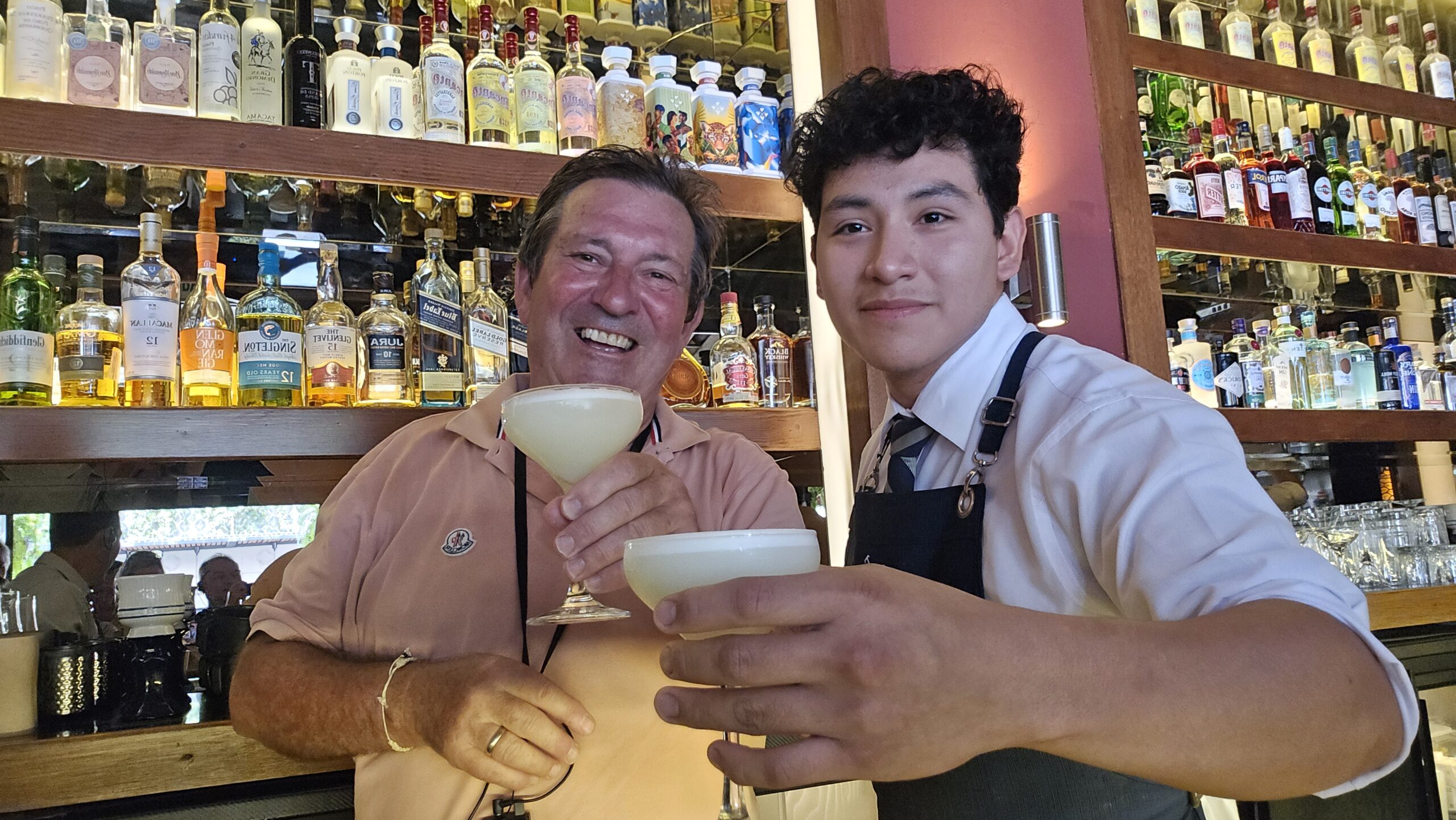 Un viajero y el bartender brindan con dos cócteles en la barra. Detrás, los estantes están llenos de botellas.