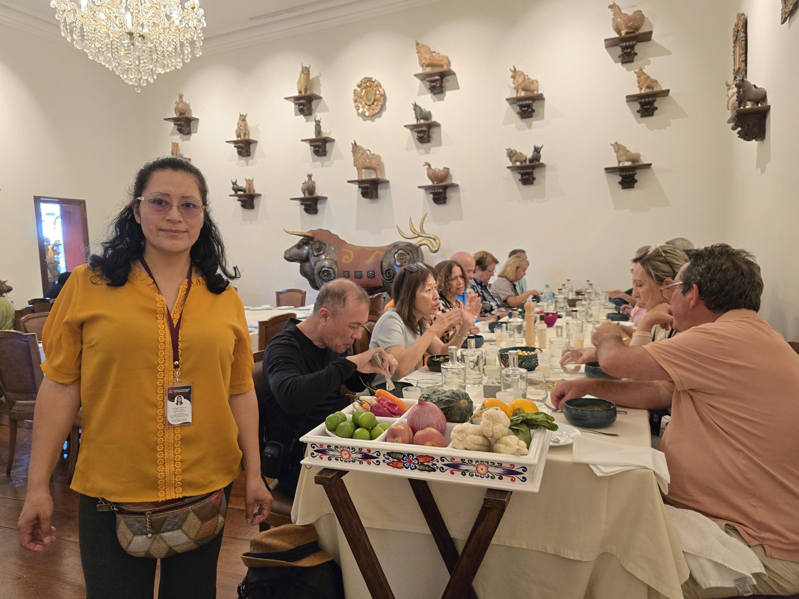 Una anfitriona presenta una bandeja con limones, ajíes, cebolla y zapallo, mientras el grupo se sienta a la mesa. En la pared hay estantes con figuras de animales.