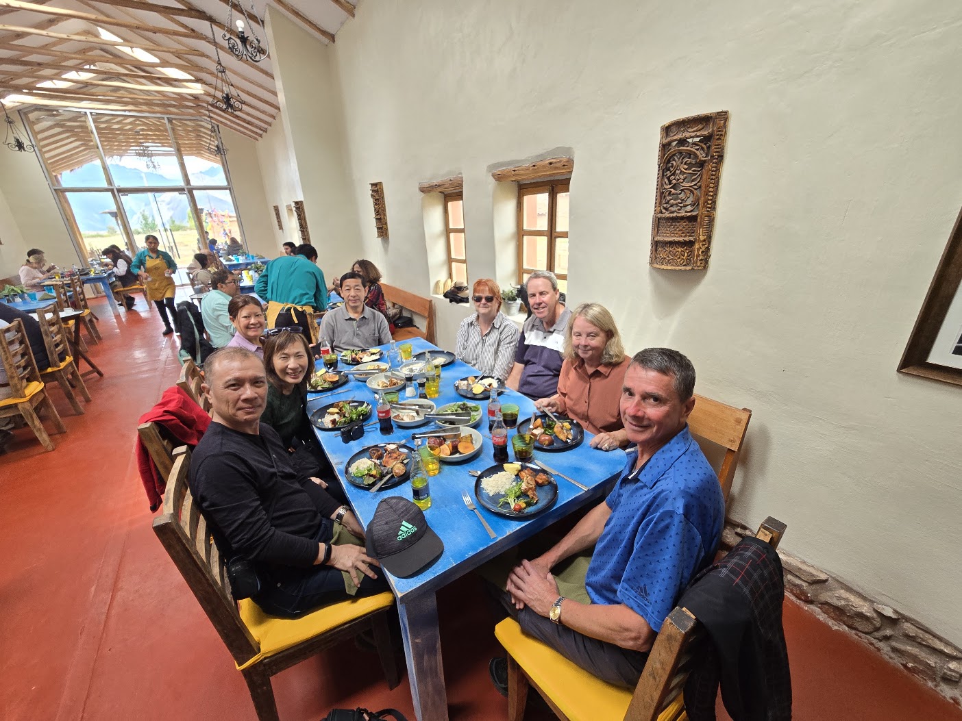 Un grupo almuerza en una mesa azul, con platos servidos y jugos. Por la ventana se ven los cerros.