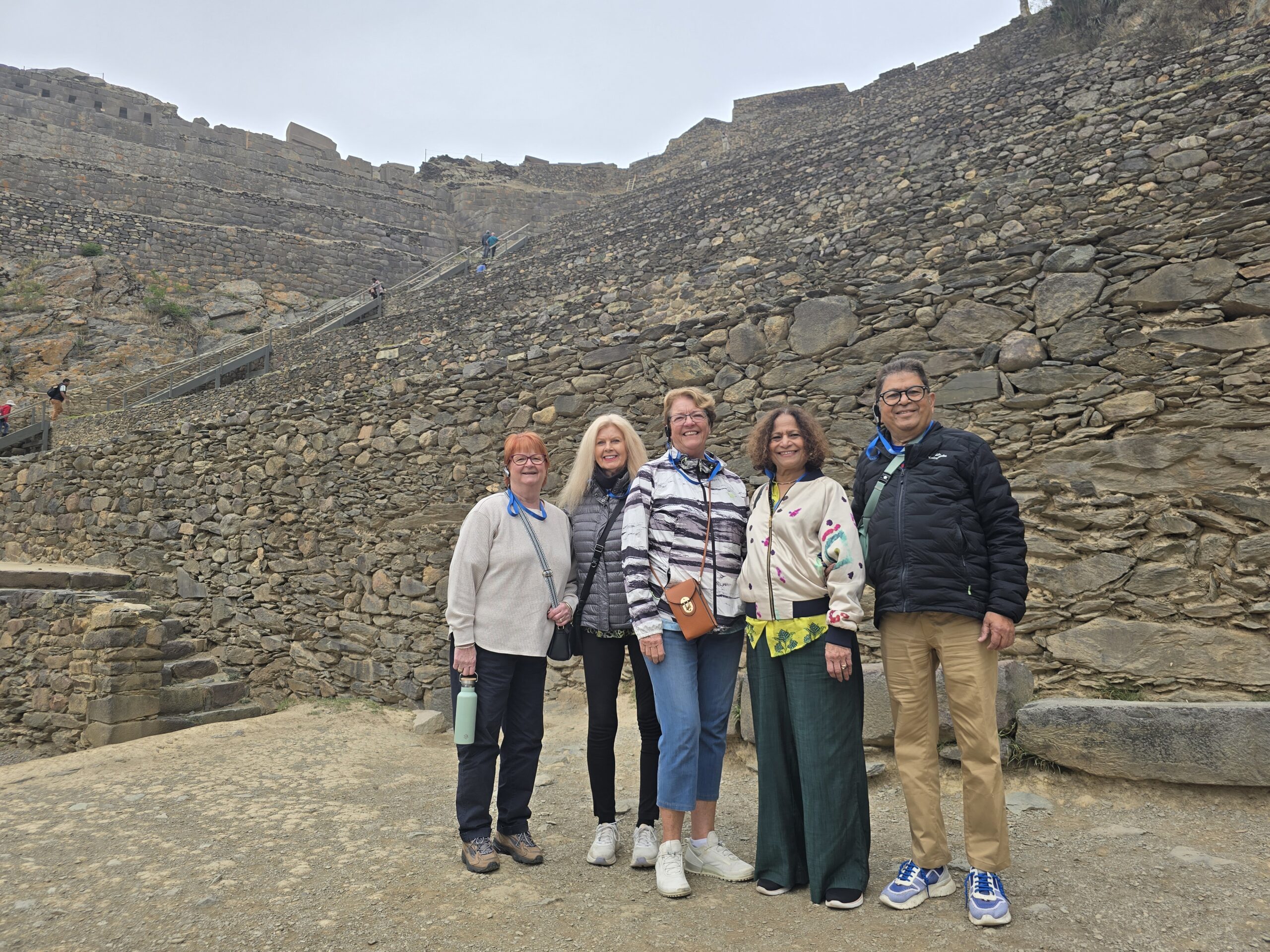 Cinco viajeros posan al pie de los andenes de piedra de Ollantaytambo. Unas escaleras suben por el muro, bajo un cielo nublado.