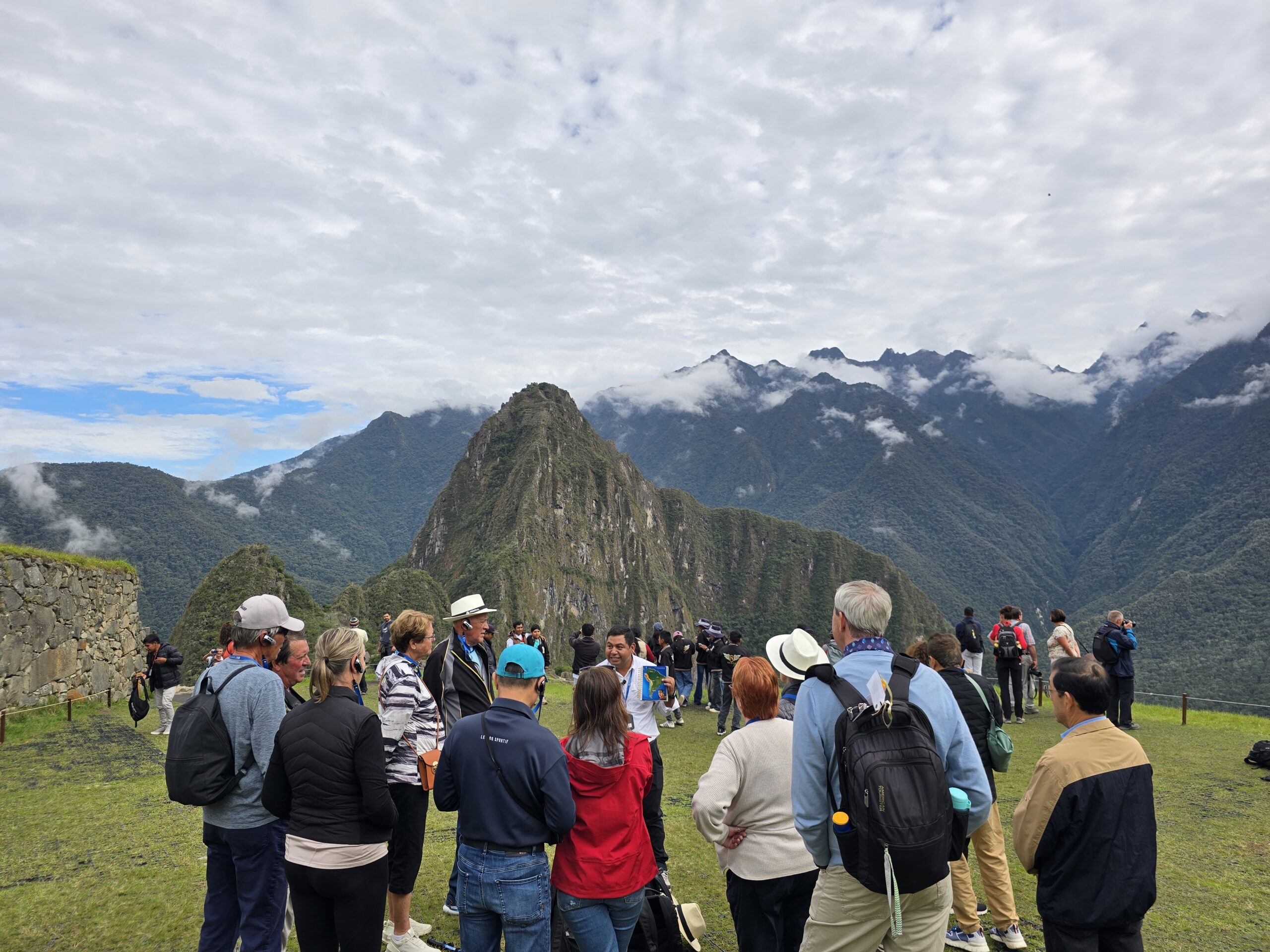 Un grupo de viajeros, de espaldas, contempla Machu Picchu. El Huayna Picchu se levanta entre nubes, sobre las montañas verdes.