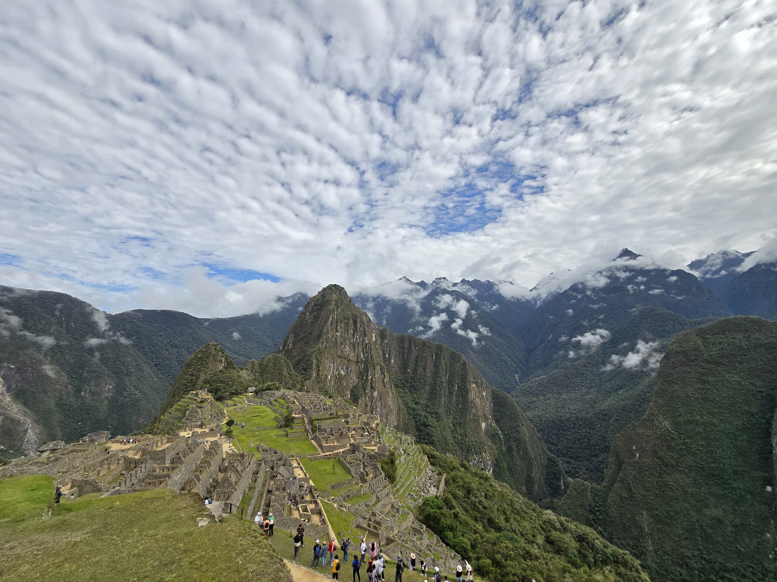 Machu Picchu, una de las Siete Maravillas del mundo moderno, aparece entre las nubes: terrazas, plazas verdes y el Huayna Picchu al fondo, con viajeros asomados al filo de la ciudadela.