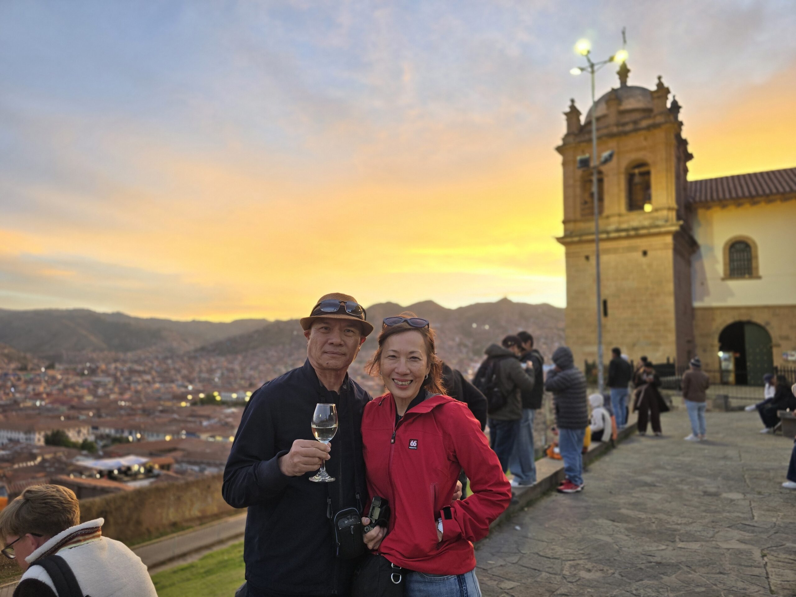 Una pareja brinda al atardecer, con el Cusco iluminado a sus pies. A un lado se ve la torre de una iglesia y el cielo en naranja.