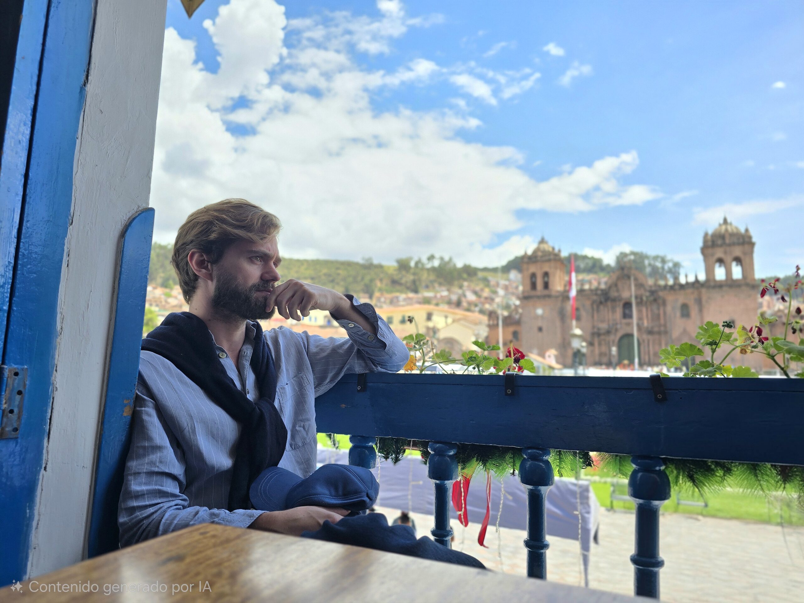 Un viajero mira desde un balcón azul hacia la Catedral del Cusco y los techos de la ciudad. El cielo está azul con nubes blancas.