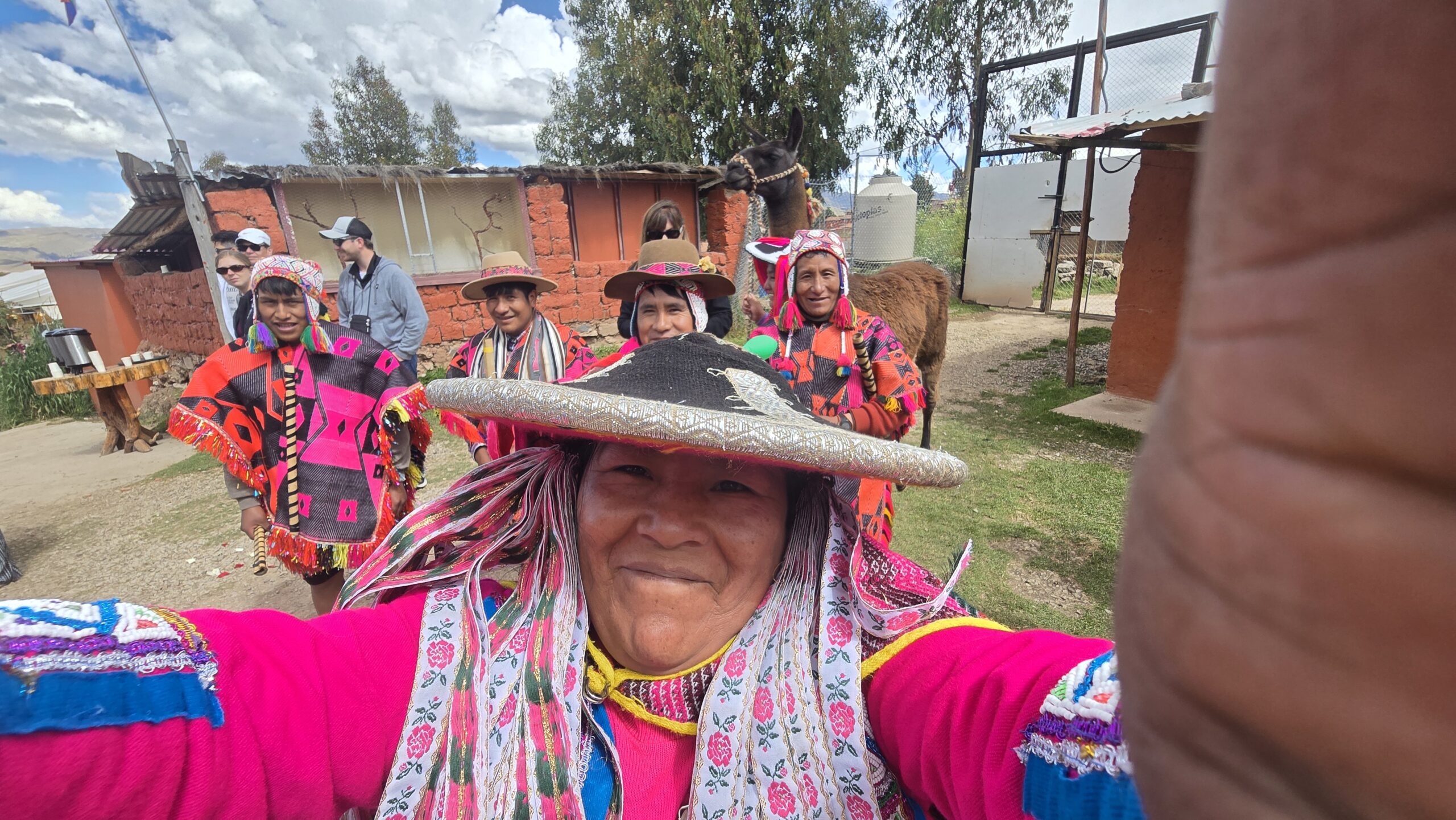 Una mujer con sombrero bordado se toma una selfie junto a su comunidad. Detrás se ve una llama y unas casas de adobe.