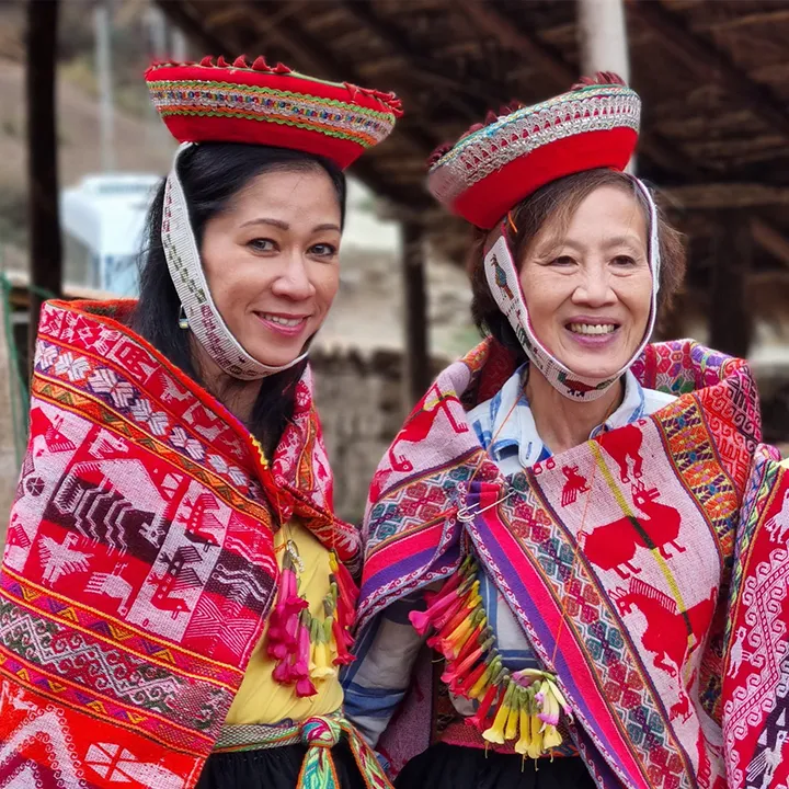 Dos mujeres sonríen muy de cerca, con monteras rojas y llicllas tejidas donde se repiten llamas y grecas de colores.
