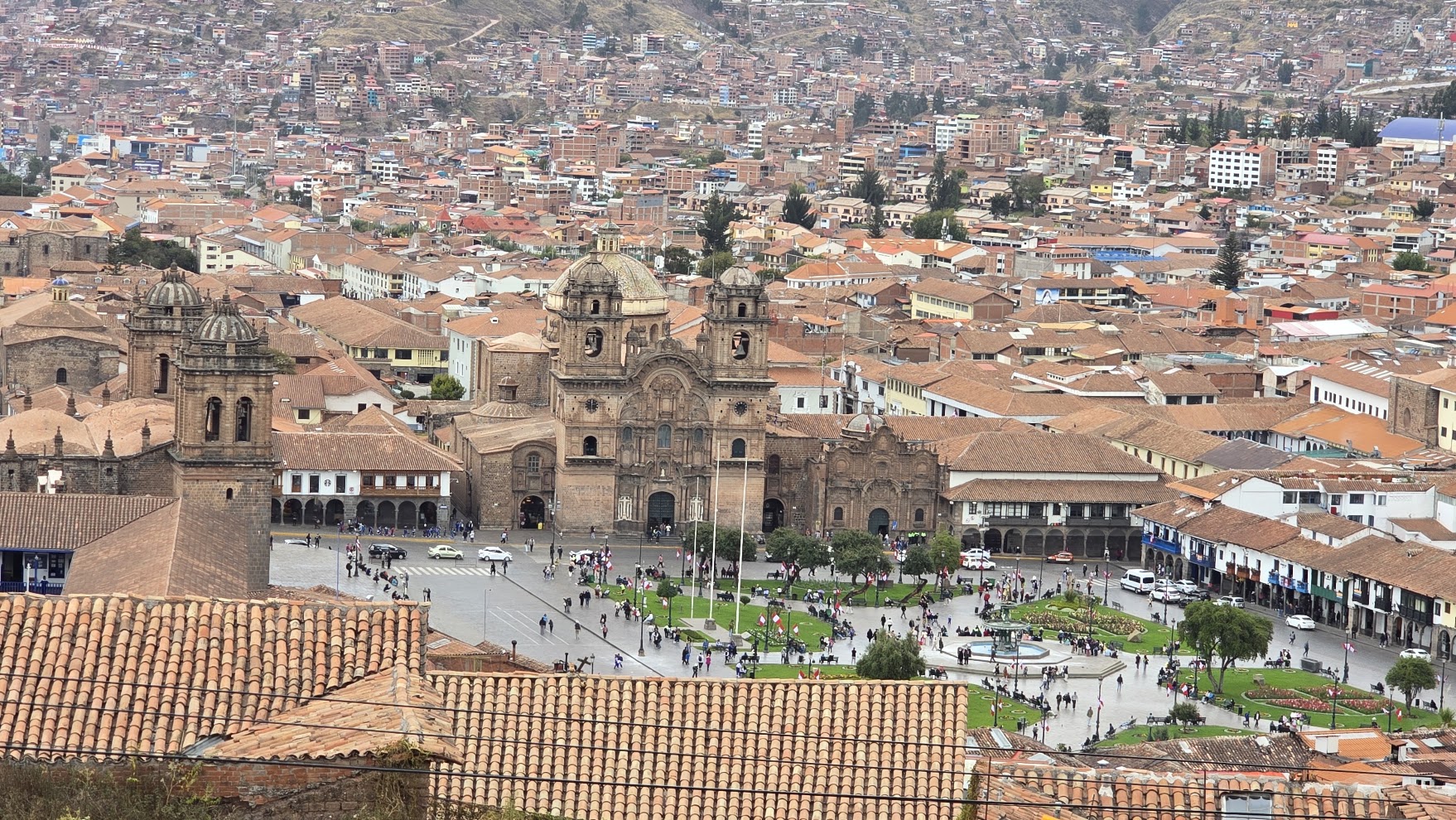 La Catedral del Cusco se ve en el centro de la Plaza de Armas, rodeada de techos de teja. La ciudad sube por los cerros y la plaza está llena de gente.