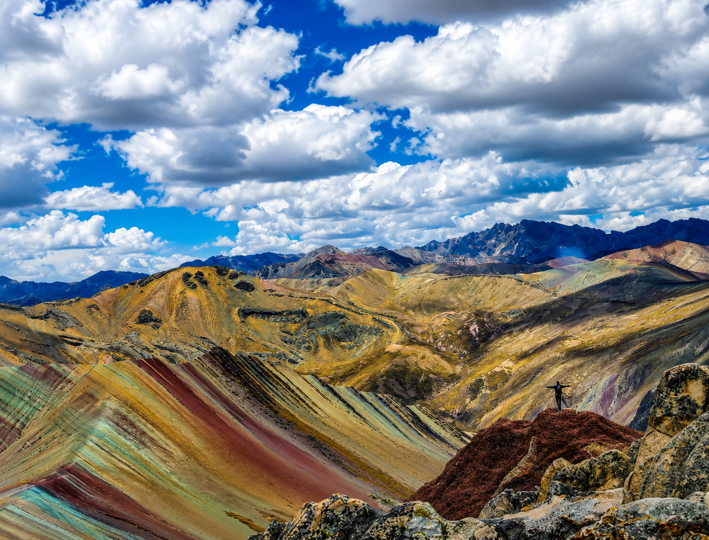 Una persona abre los brazos sobre una roca, frente a las franjas de la Montaña de Colores. El cielo está azul y lleno de nubes.