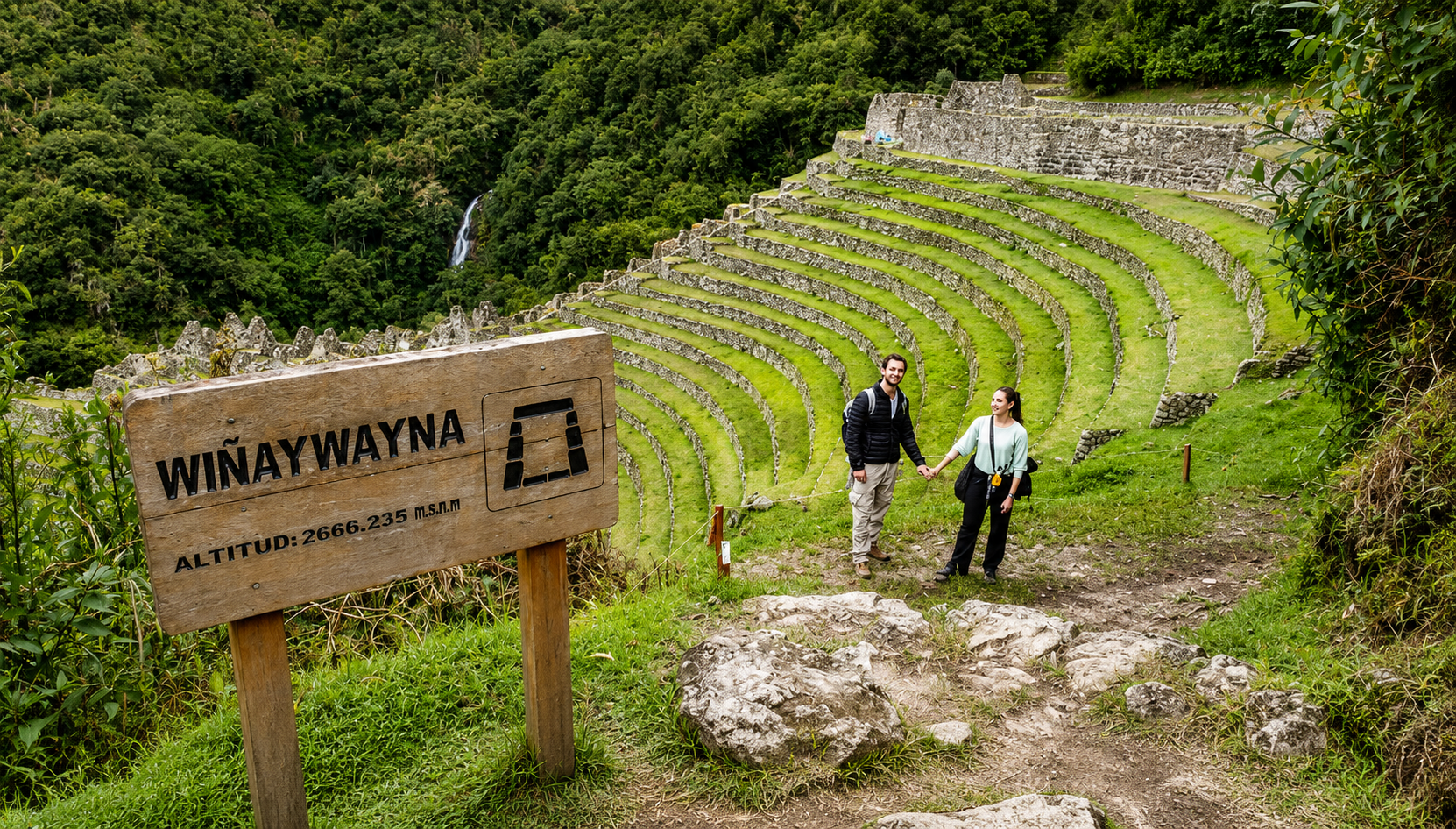 Una pareja se toma de la mano frente a las terrazas curvas de Wiñay Wayna. Al fondo, la selva cubre la ladera y se ve una cascada.