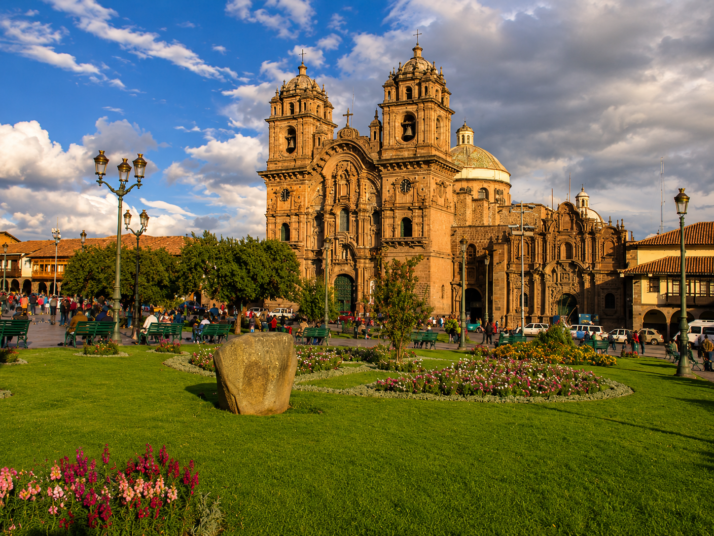 La Catedral del Cusco se ve dorada por el sol de la tarde, detrás de los jardines de la plaza. Hay bancas, faroles y gente paseando bajo un cielo con nubes.