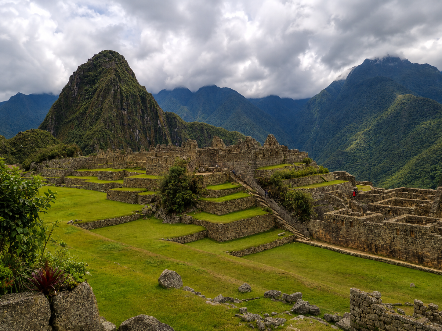 Los andenes de Machu Picchu se ven verdes bajo un cielo de nubes, con el Huayna Picchu al fondo. Unas escaleras de piedra suben entre los muros de la ciudadela.