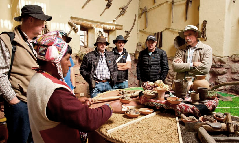 En el interior de una casa de adobe, un anfitrión con chullo explica las papas nativas. Los viajeros rodean la mesa, donde se alinean cuencos, mazorcas y cerámica.