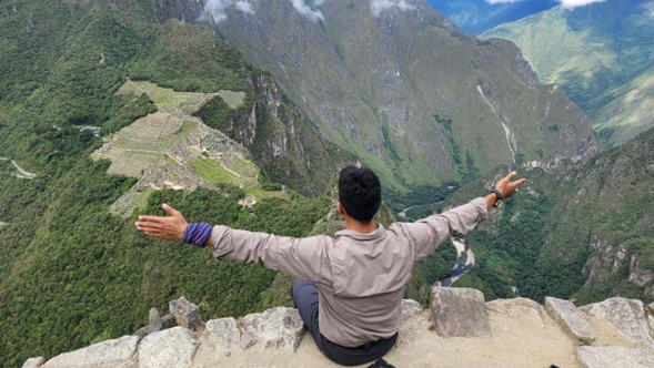 Un viajero abre los brazos sentado en una piedra, con Machu Picchu abajo en el valle. Alrededor se ven montañas verdes y el río.