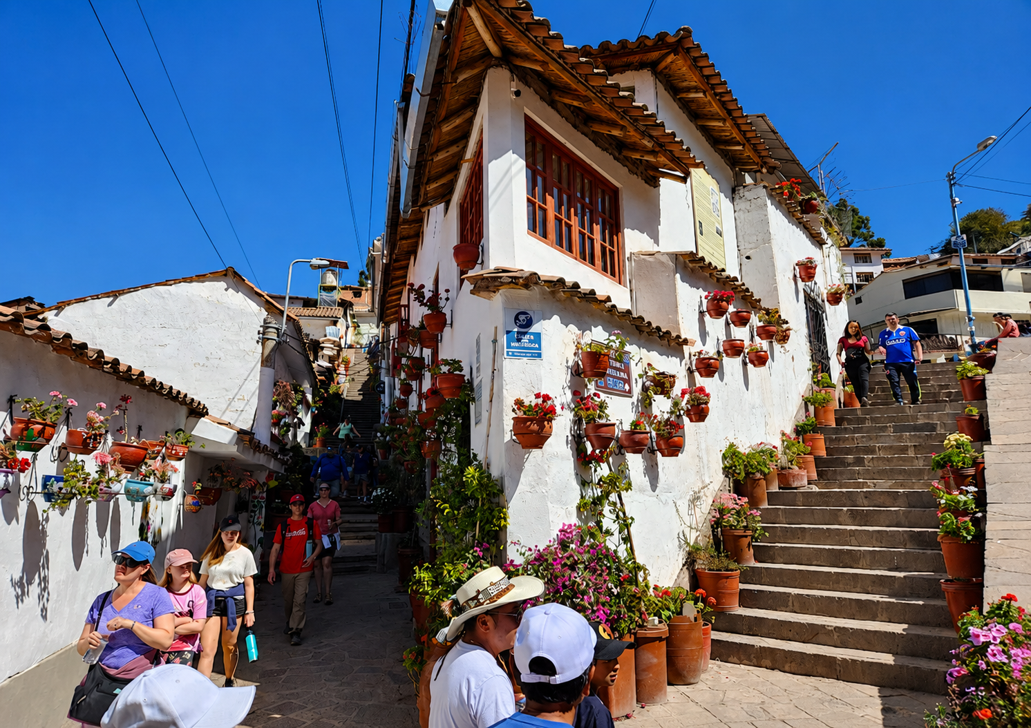 Un callejón de paredes blancas, lleno de macetas con flores, en el Monasterio de Santa Catalina. La gente sube las escaleras bajo un cielo limpio.