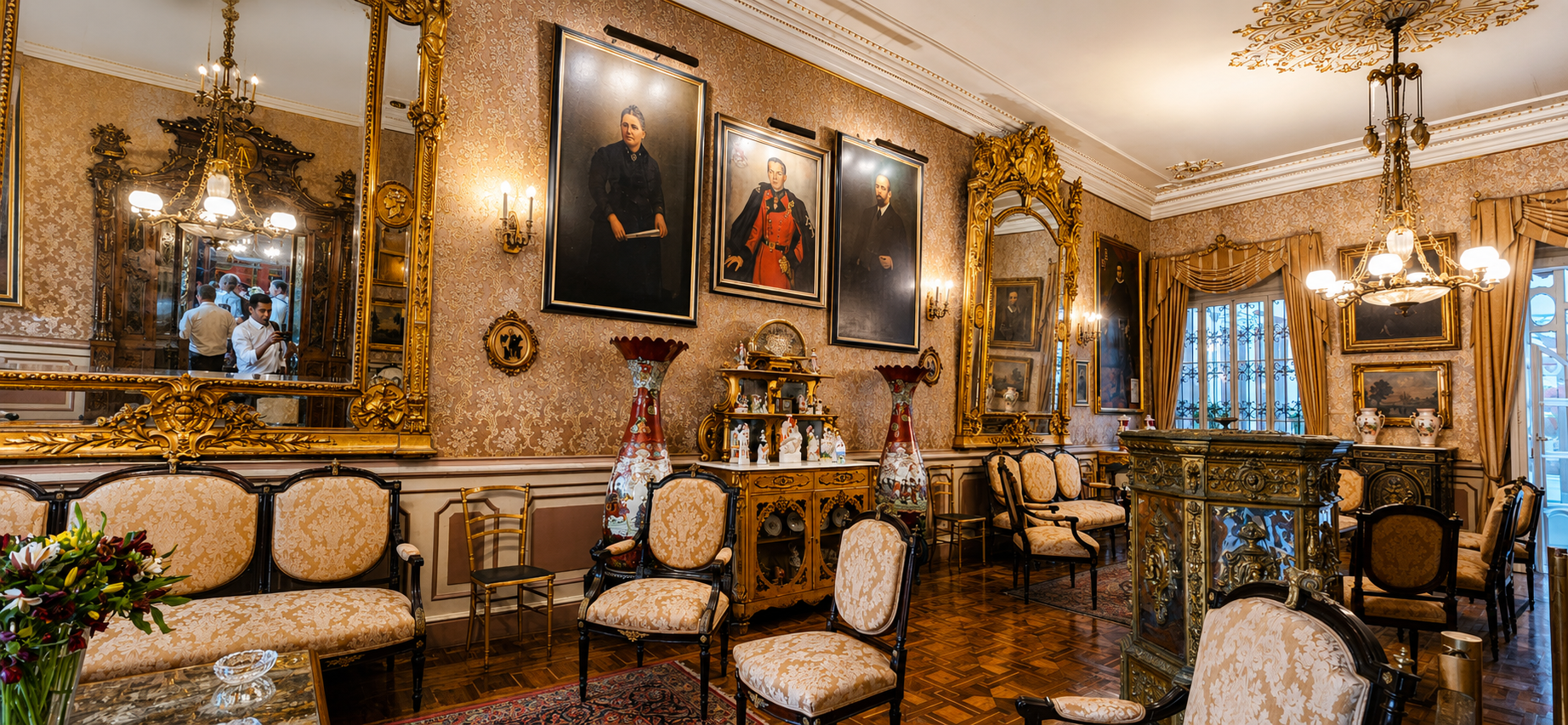 Un salón de espejos dorados, retratos antiguos y lámparas de araña. Sofás y sillas de época rodean el piso de madera.