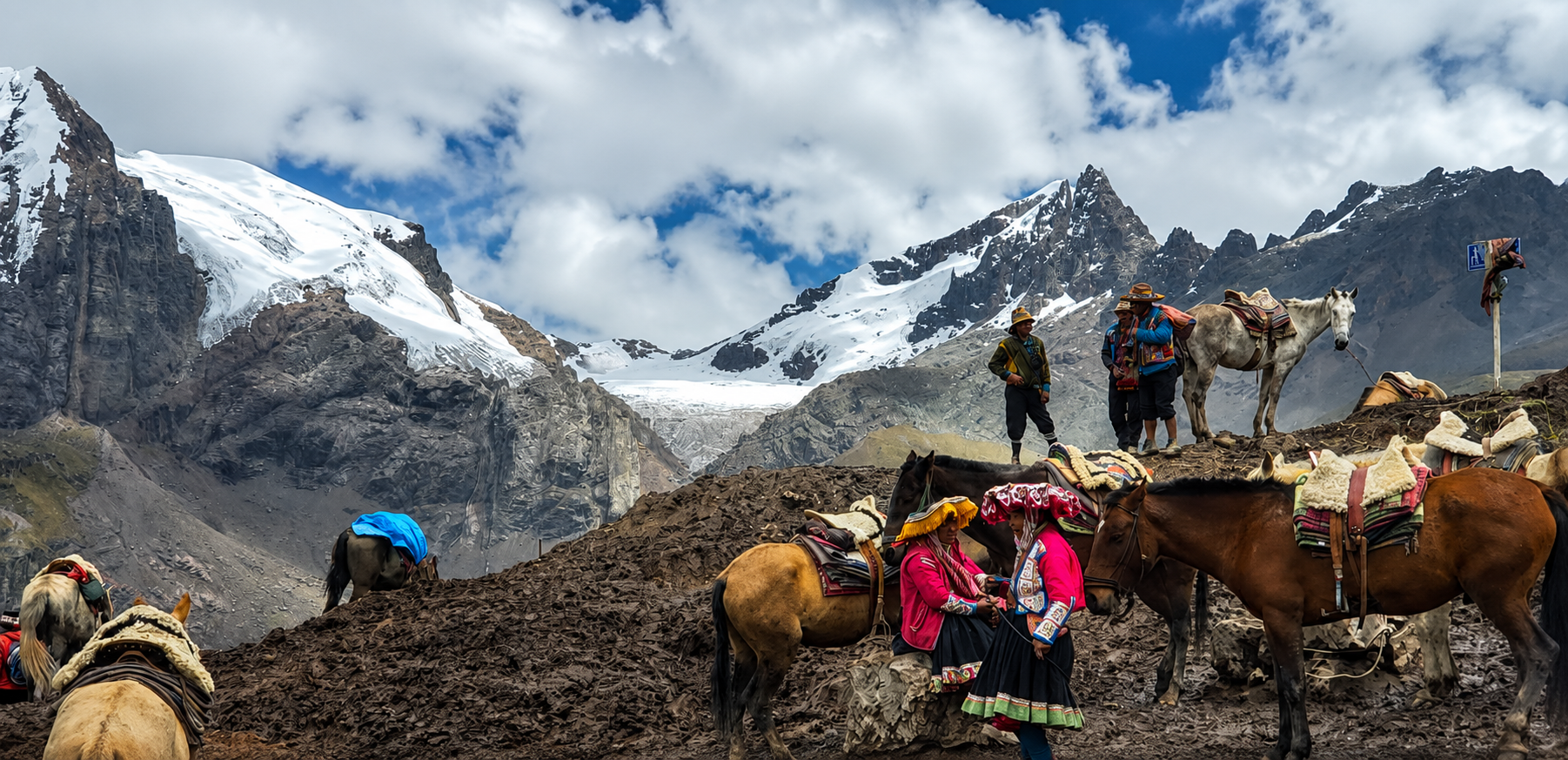 Personas con traje tradicional esperan junto a caballos ensillados en un paso alto de los Andes. Detrás, nevados y un glaciar bajo un cielo de nubes.