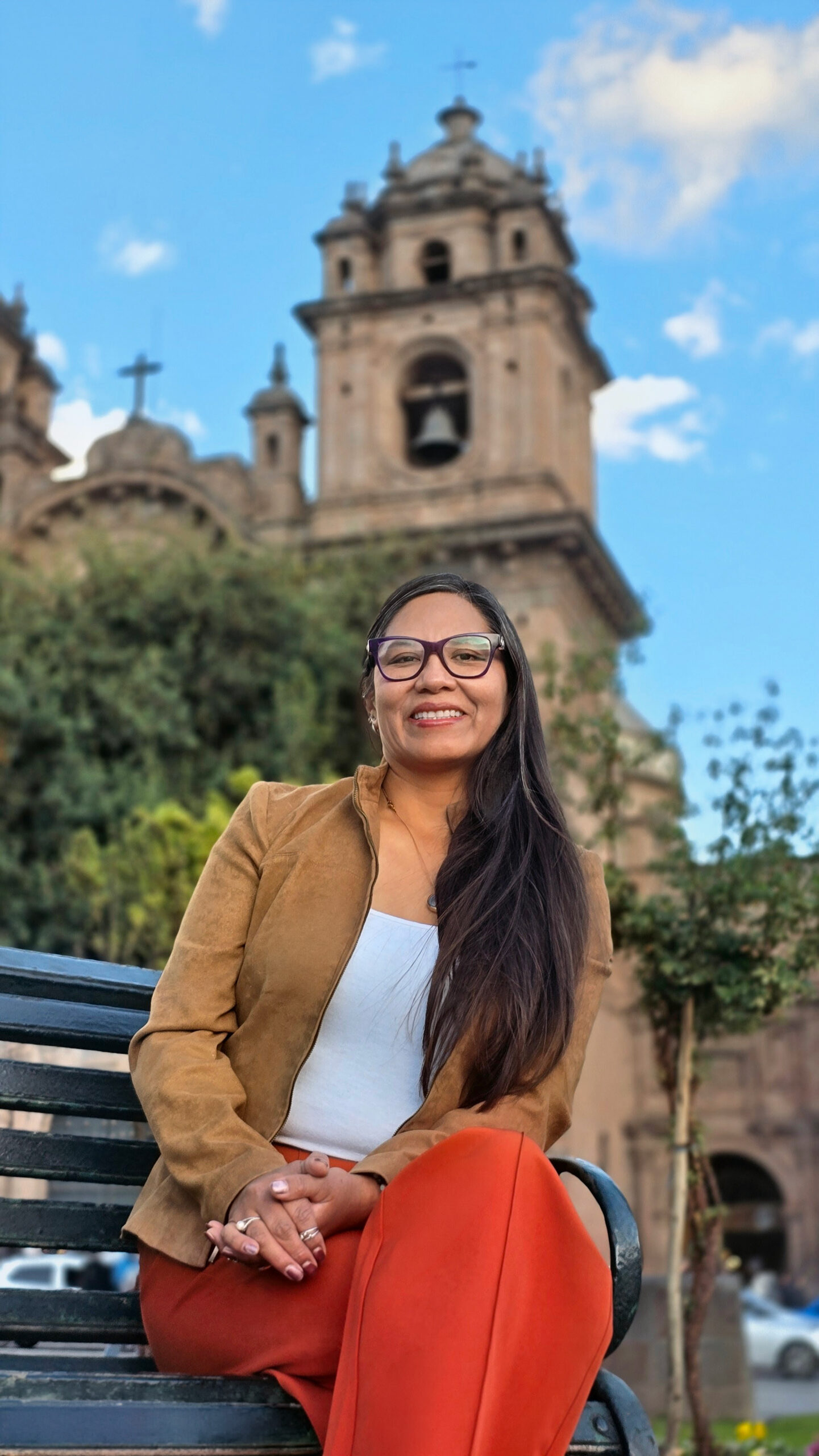 La fundadora de InkaTraces, que esta vestida de chaqueta camel y pantalón rojo sonríe sentada en una banca. Detrás, la torre de una iglesia colonial del Cusco.