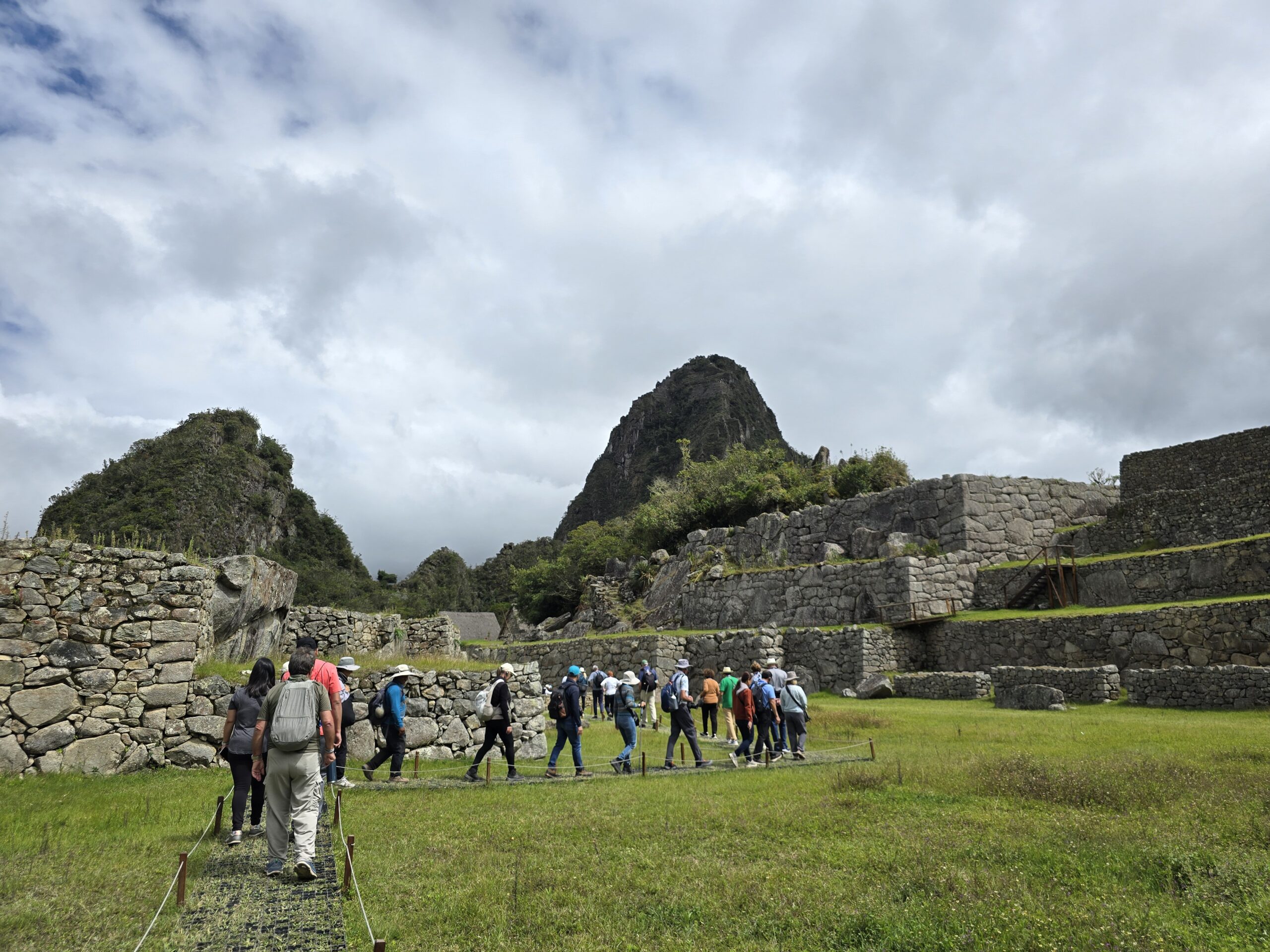 Un grupo de visitantes camina entre los muros de piedra de Machu Picchu. Al fondo se ve el Huayna Picchu, bajo un cielo nublado.