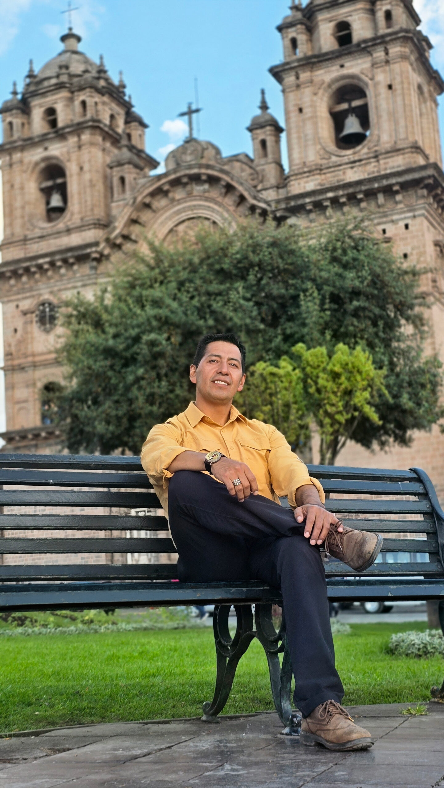 El fundador de InkaTraces vestido de camisa amarilla, descansa en una banca de la Plaza de Armas del Cusco, con la Catedral y sus dos torres detrás.