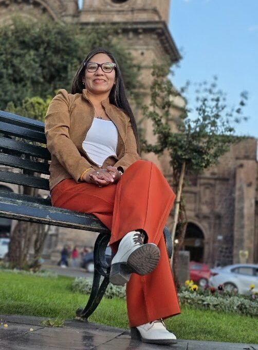 La fundadora de InkaTraces, que esta vestida de chaqueta camel y pantalón rojo sonríe sentada en una banca. Detrás, la torre de una iglesia colonial del Cusco.