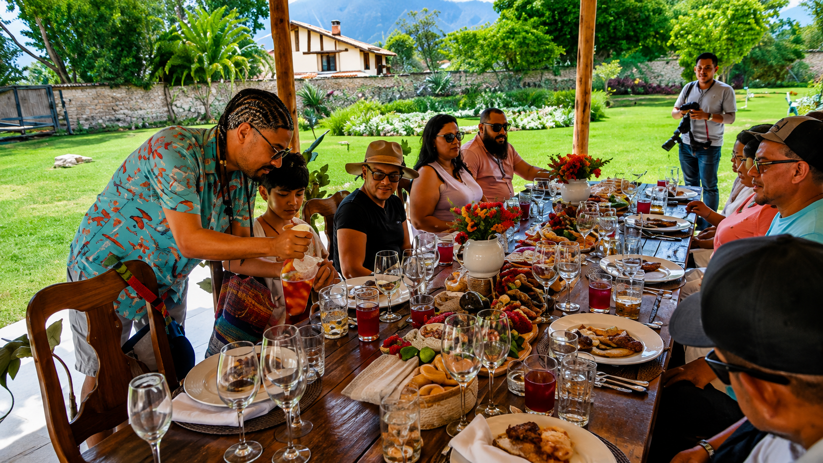 Un hombre sirve un vaso en una mesa larga, llena de panes, frutas y platos, en un jardín. Detrás se ve el césped, una casa y los cerros.