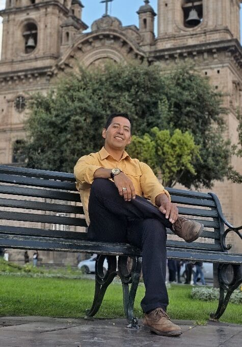 El fundador de InkaTraces vestido de camisa amarilla, descansa en una banca de la Plaza de Armas del Cusco, con la Catedral y sus dos torres detrás.