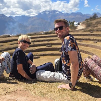 Un padre y su hijo se sientan al sol frente a los andenes circulares de Moray. Al fondo, la cordillera nevada cierra el Valle Sagrado