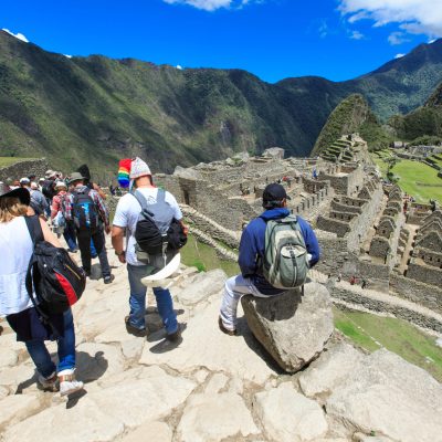 Viajeros avanzan por los senderos de piedra de Machu Picchu, una de las Siete Maravillas del mundo moderno. Uno se sienta en una roca a mirar las terrazas y la ciudadela.